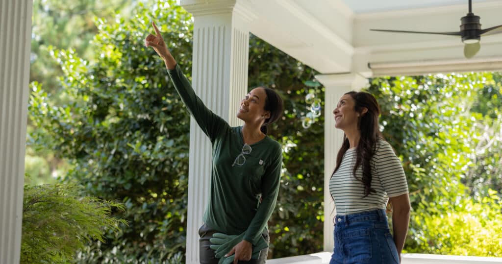 Aptive Pest Control Specialist Pointing Out Potential Pest Entry Points To A Smiling Homeowner On A Porch Surrounded By Greenery.