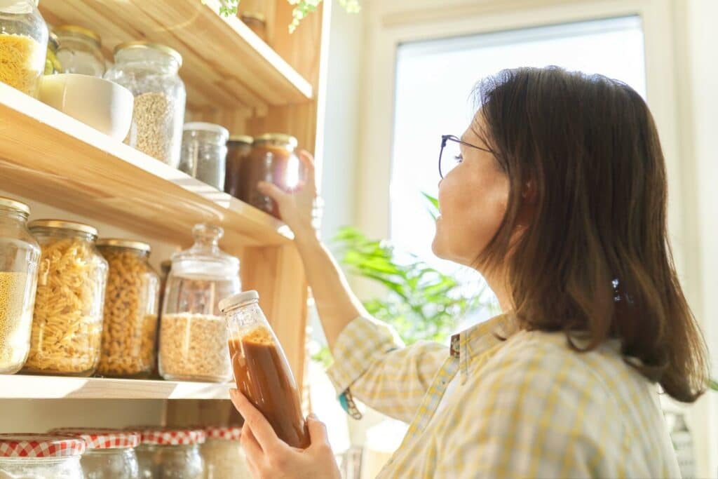 A Woman With Glasses And Long Dark Hair In A Yellow Plaid Shirt Reaching For A Jar On A Wooden Pantry Shelf, Surrounded By Jars Filled With Various Dry Foods Like Pasta And Grains, With A Bright Window In The Background.