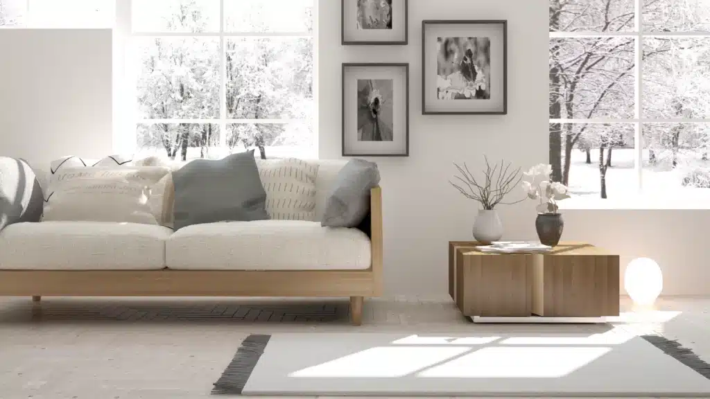 A Cozy Living Room Interior With A Light Beige Sofa Adorned With Various Pillows, A Wooden Coffee Table With Decorative Vases, And Large Windows Showcasing A Snowy Outdoor Scene With Trees Covered In Snow.