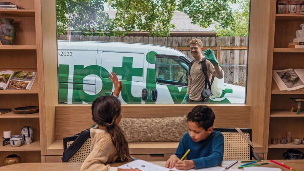 An Aptive Pest Control Specialist Waves To Two Children From Outside A House Window, With An Aptive Van Parked In The Background. The Children Are Sitting At A Table Inside, One Waving Back While The Other Is Drawing.