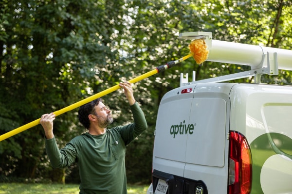 An Aptive Pest Control Specialist Uses A Long Yellow Pole To Clean The Roof Of A Company Van.