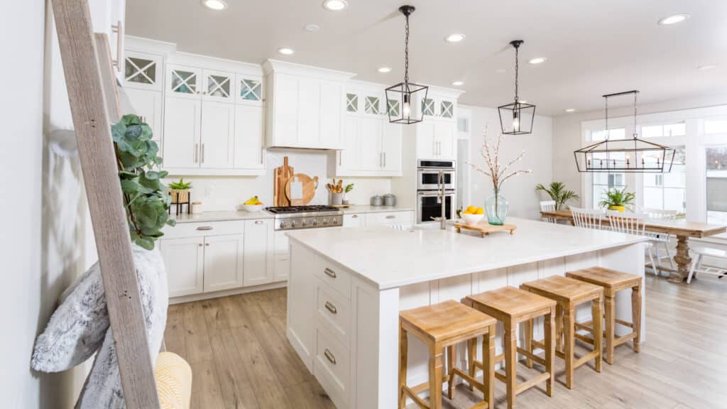 Bright And Modern Kitchen With White Cabinets, A Large Central Island With Wooden Stools, And Stainless Steel Appliances. The Room Is Decorated With Fresh Plants And Natural Light Floods In Through Large Windows, Creating A Welcoming And Airy Atmosphere.