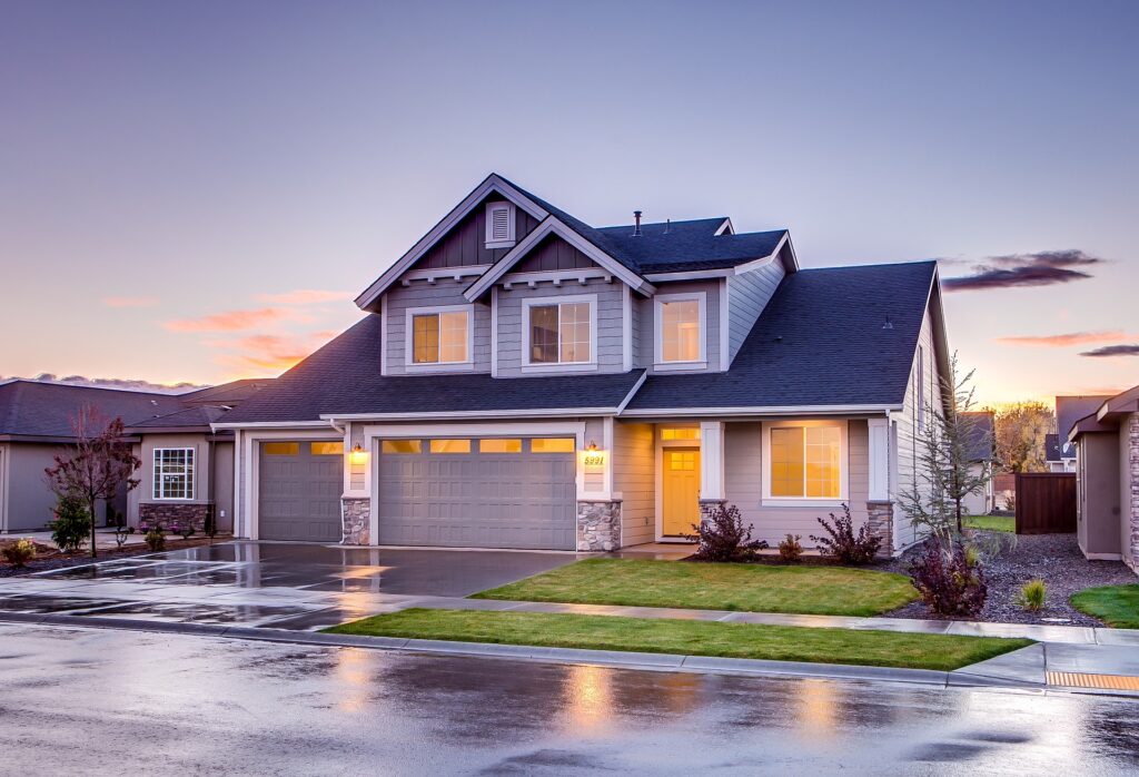 A Modern Two-Story Suburban Home With Gray Siding And A Dark Roof, Illuminated By Warm Interior Lights During Sunset. The House Features A Large Three-Car Garage, Well-Manicured Lawn, And A Driveway With Reflections From Recent Rain. The Sky Is Tinged With Soft Hues Of Pink And Purple As The Day Transitions Into Evening.