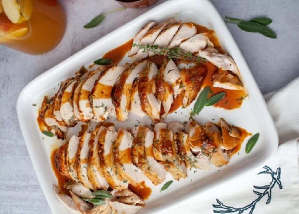 Sliced Apple Cider-Glazed Turkey Breast Arranged On A White Platter, Garnished With Fresh Sage Leaves And Thyme, With A Side Of Iced Apple Cider.