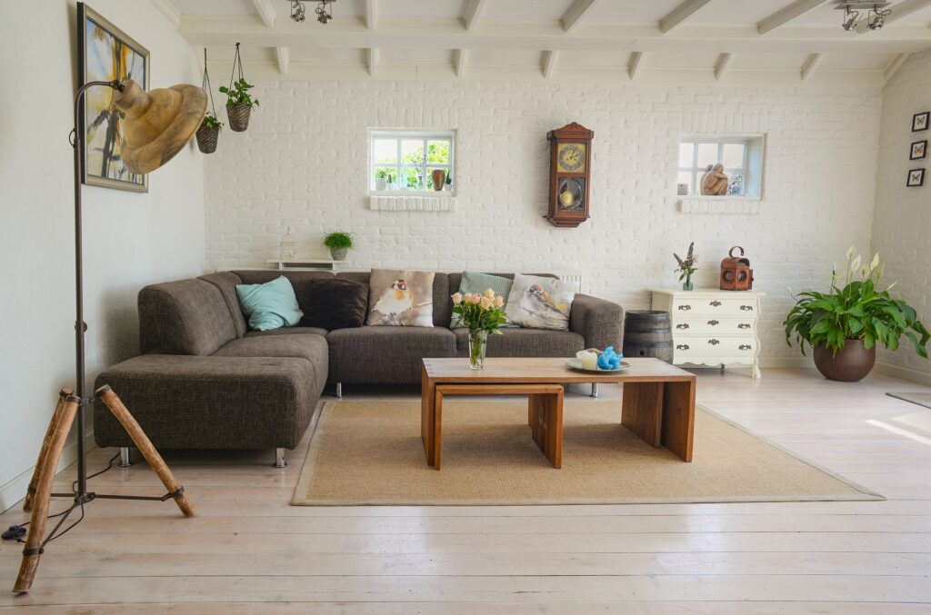 A Cozy Living Room Interior With A Brown Sectional Sofa Adorned With Decorative Bird-Themed Pillows, A Small Coffee Table With Flowers, And A Potted Plant In The Corner. The Room Features A White Brick Wall With Hanging Plants And A Vintage Clock.