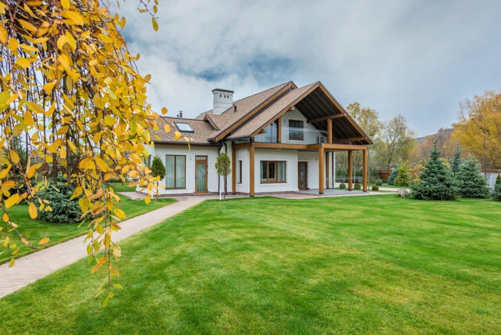 A Modern Suburban House With A Wooden Roof And Large Windows, Surrounded By A Well-Maintained Green Lawn And Autumn Trees With Yellow Leaves.