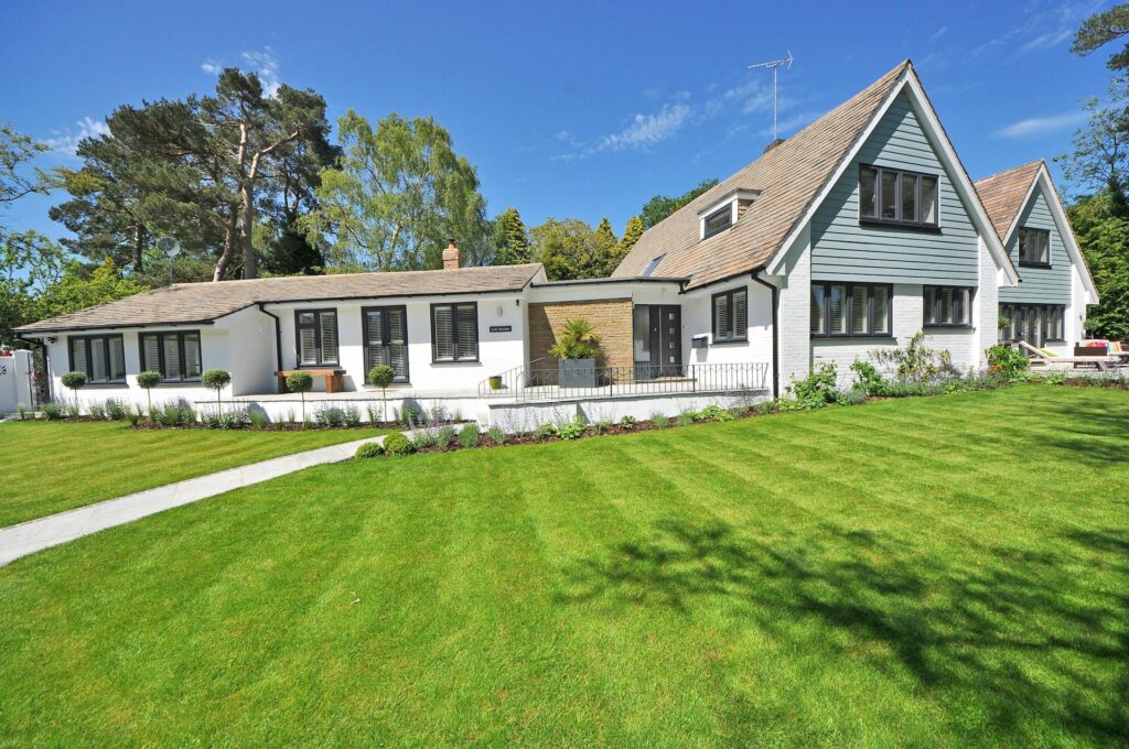 A Modern White House With A Green Roof And Large Windows, Surrounded By A Well-Manicured Lawn And Garden, Set Against A Blue Sky And Trees.