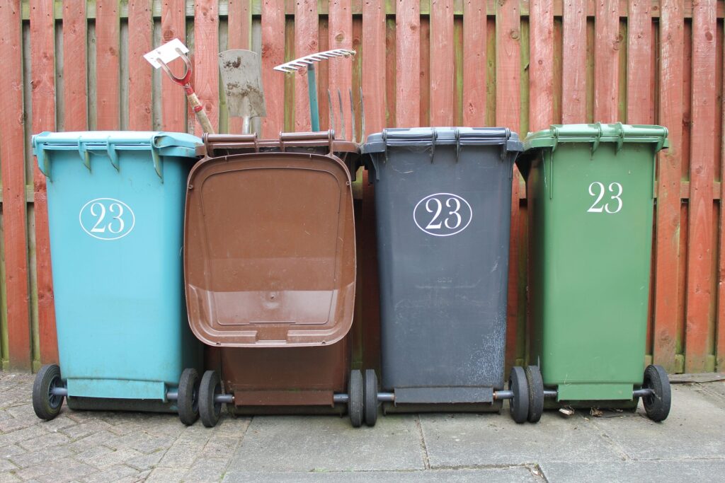 Four Outdoor Garbage Bins In Blue, Brown, Gray, And Green Placed Side By Side, Marked With The Number 23. The Bins Are Against A Red Wooden Fence With Gardening Tools Resting Inside One Of The Bins.